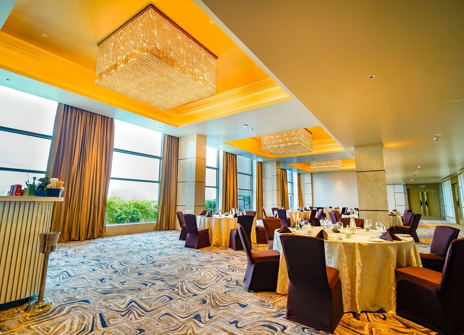 A spacious banquet hall with round tables, elegant chandeliers, and natural light from large windows