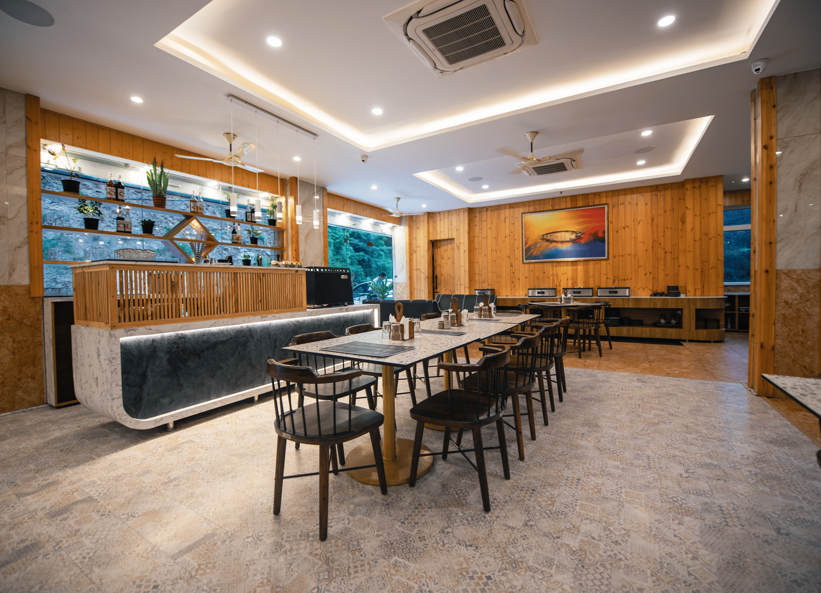 A restaurant at The Tattva Devaprayag with a long wooden table with black chairs and a bar.