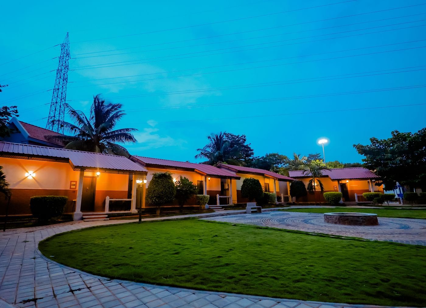 Exterior view of cottages with red roofs and green lawns at Nirantara Resort & Spa, Bengaluru, at dusk with lights illuminating the pathways.