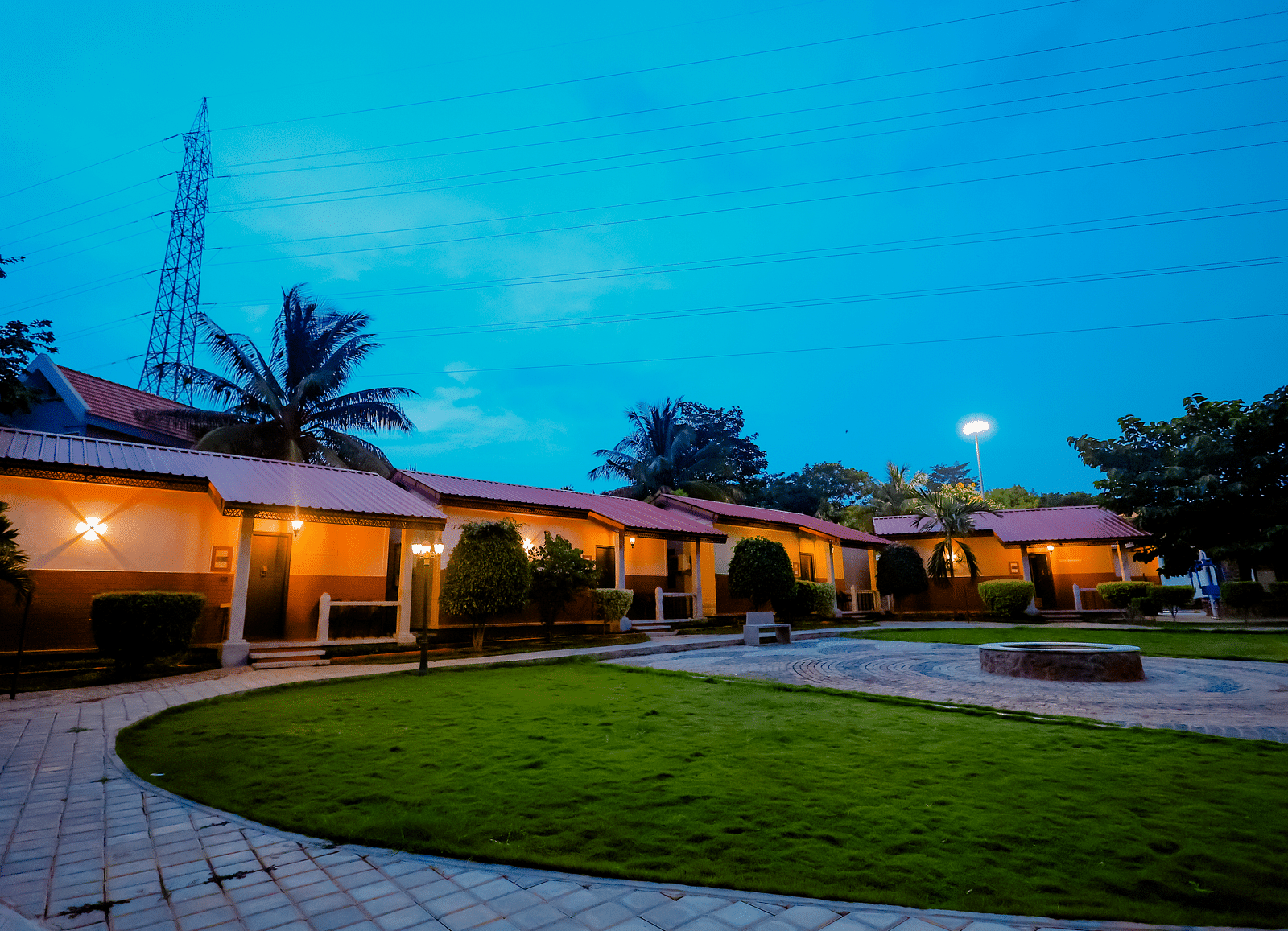 Exterior view of cottages with red roofs and green lawns at Nirantara Resort & Spa, Bengaluru, at dusk with lights illuminating the pathways.