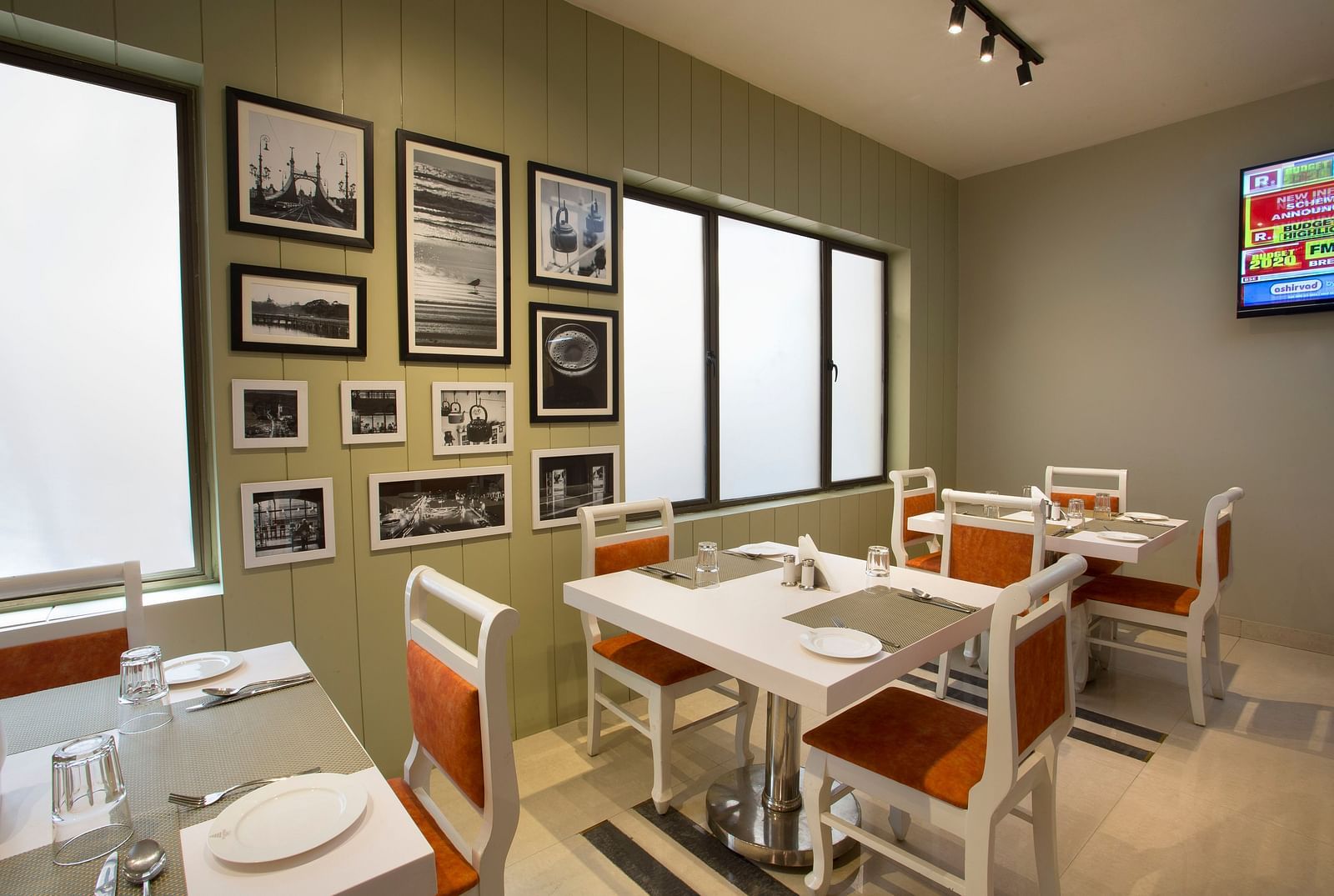 A neat dining room with white tables, orange chairs, framed black and white pictures and a TV - Hotel Langford Keys by TGI, Shanti Nagar