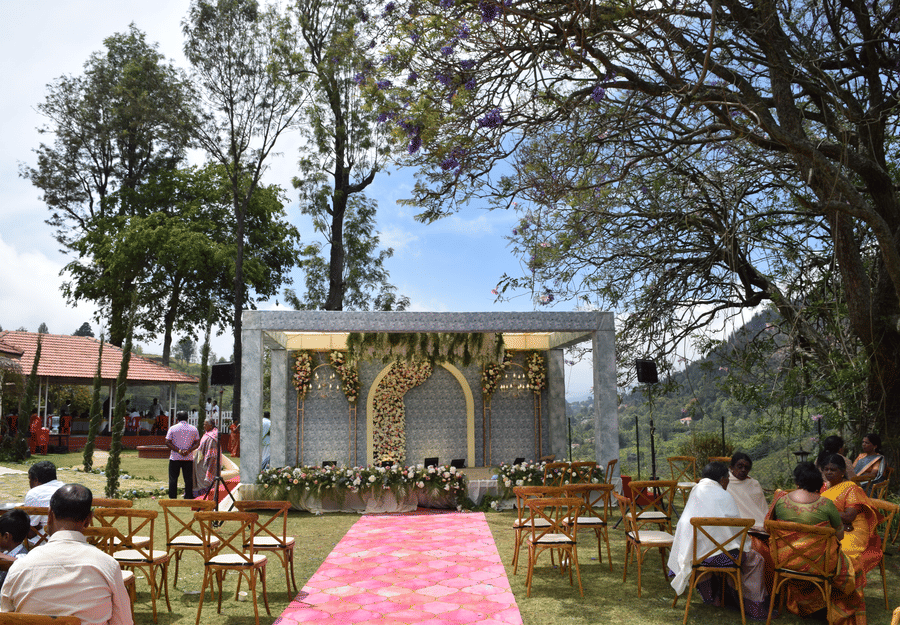 An outdoor wedding setup on a green lawn with a pink aisle runner leading to a decorated stage, surrounded by wooden chairs, with guests seated and trees in the background under a partly cloudy sky.