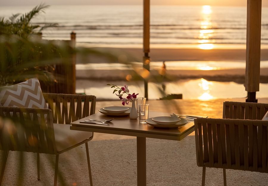a close up shot of a table with two chairs at saz on the beach restaurant during sunset with sun rays reflecting on the sea waves in the background
