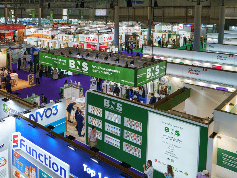 Spacious exhibition venue with rows of trade show booths, networking areas, and attendees walking through the event floor.