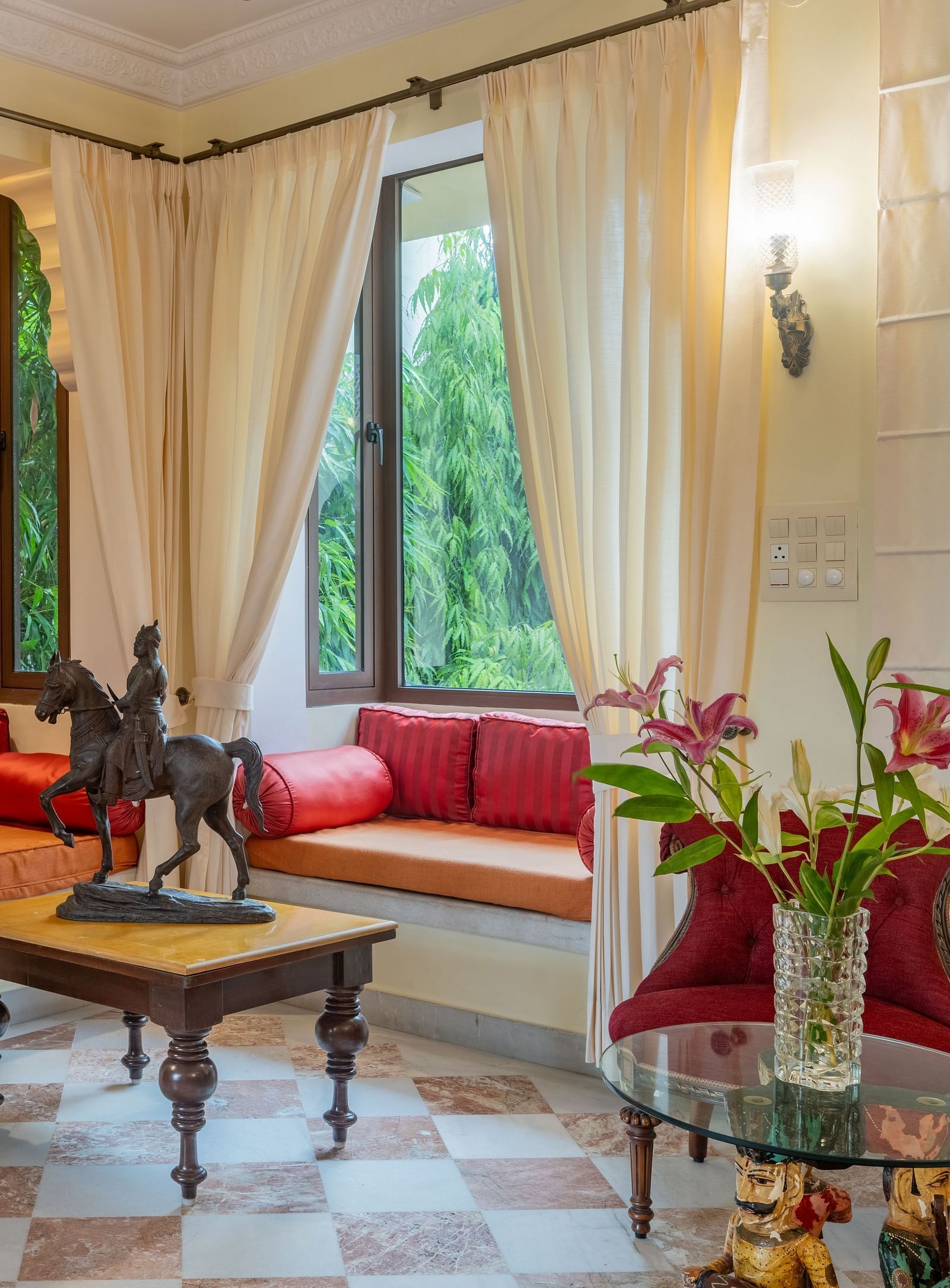 A charming sitting room at Dera Rawatsar features window seats with cushions, a wooden coffee table with a statue, and a glass-topped side table with flowers.
