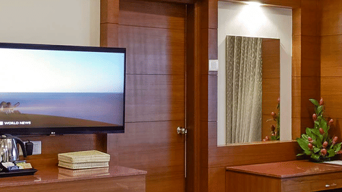 A well-lit suite at JP Hotel Chennai featuring a plush bed next to a centre table facing the TV  and a large window on its side