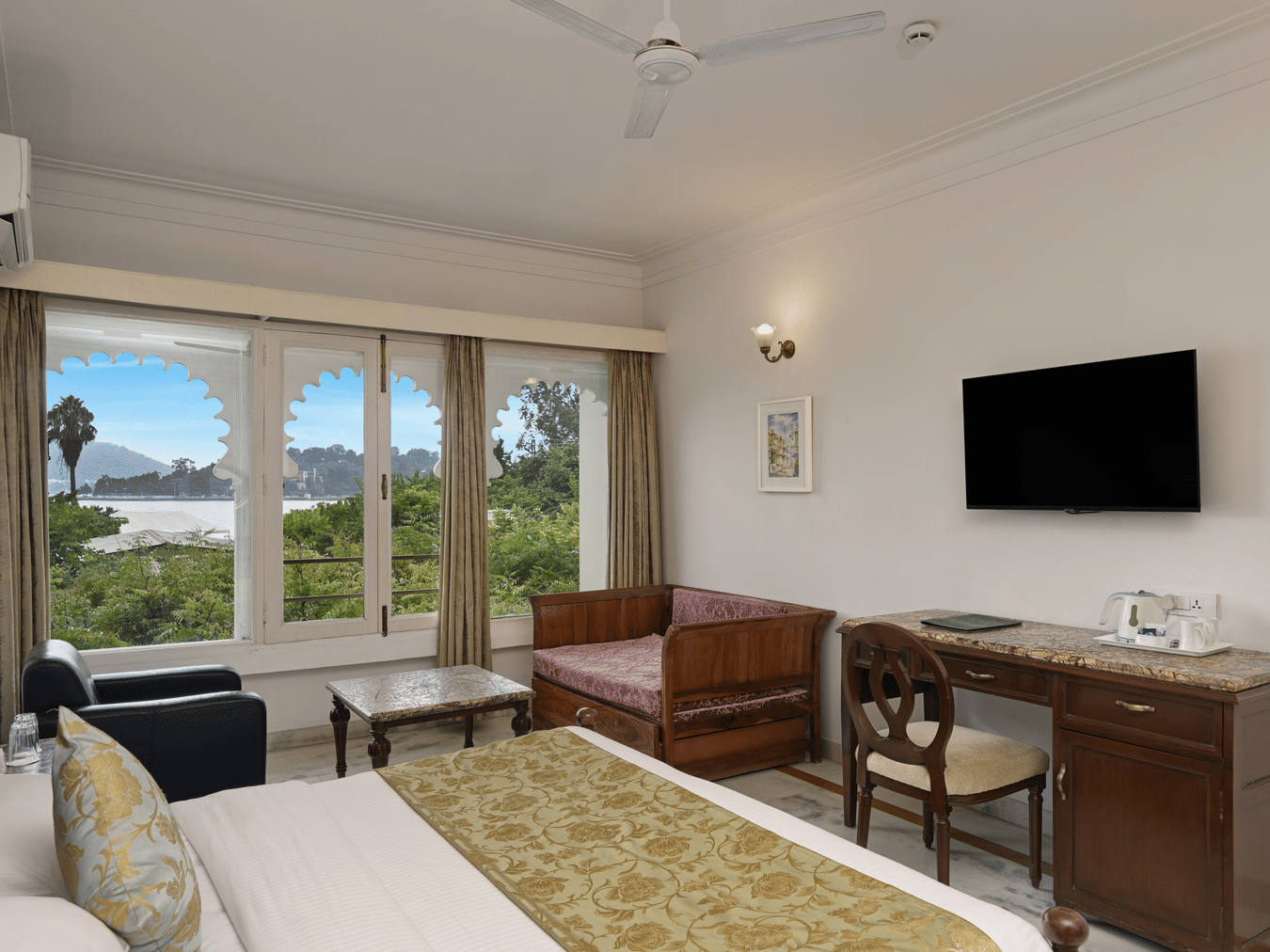 A guest room at Ram Pratap Palace with a bed, pillows, two chairs, a coffee table, a desk, a television, a lamp, and a window with arched frames overlooking the lake.