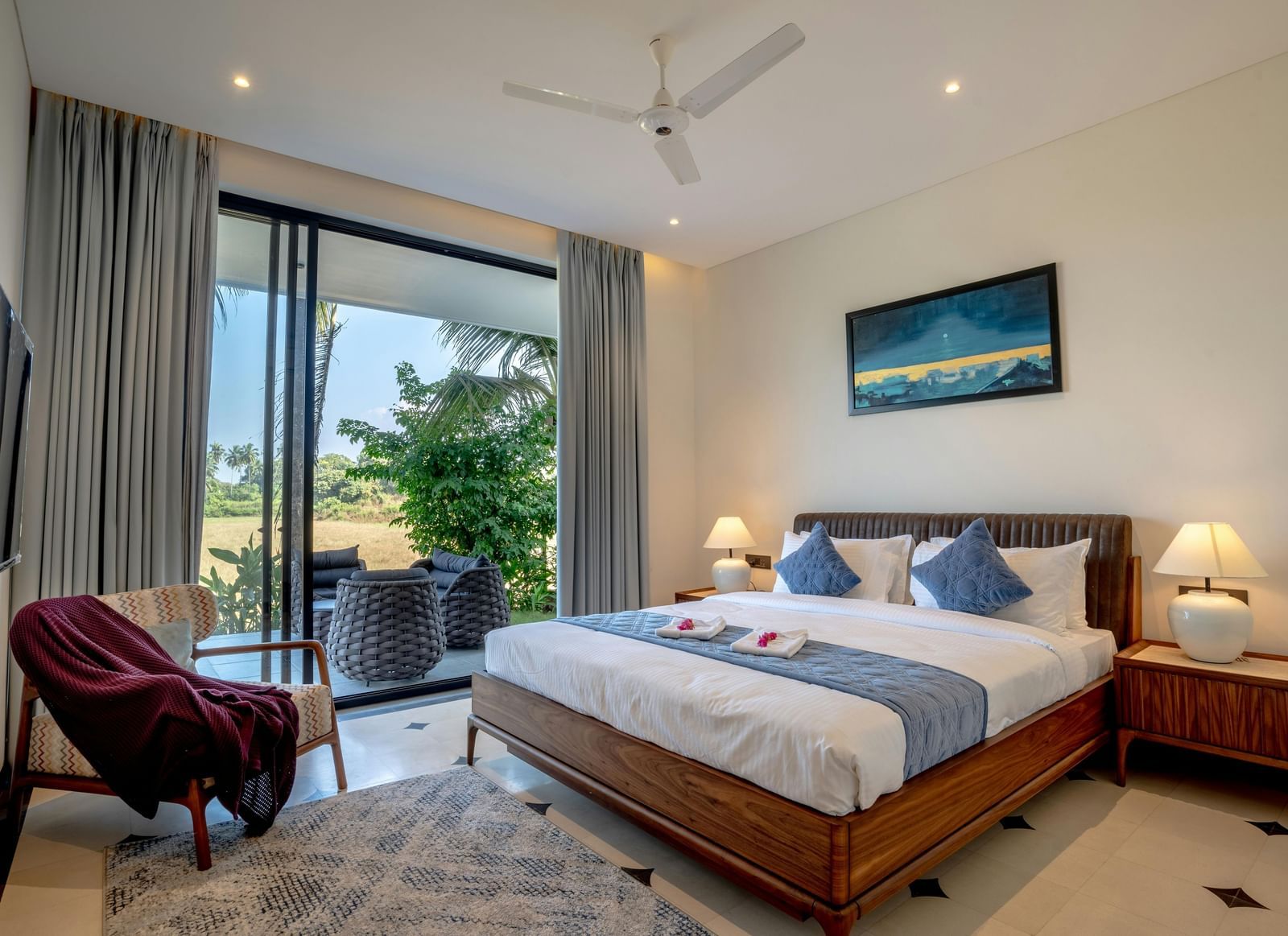 A room with balcony, spacious bed with bedside tables and lamps, artwork above the headboard, and outdoor view through glass doors