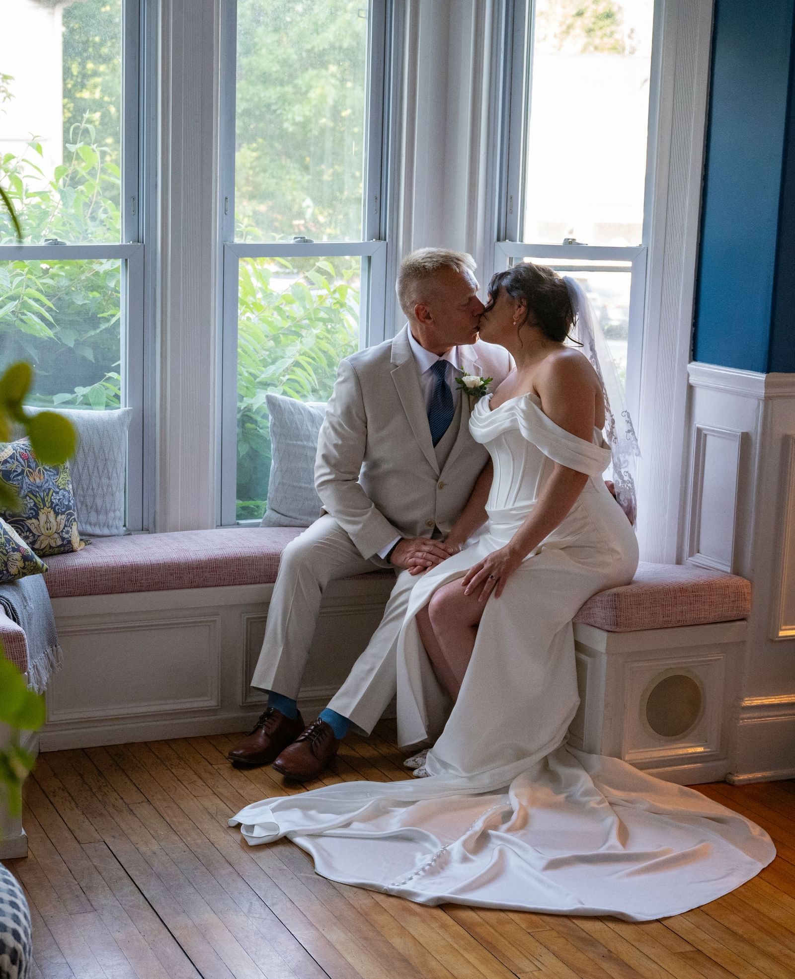 Couple in wedding attire sitting on a window seat and kissing inside Hartstone Inn, with a lit lamp, green plant, and pillows visible in the room.