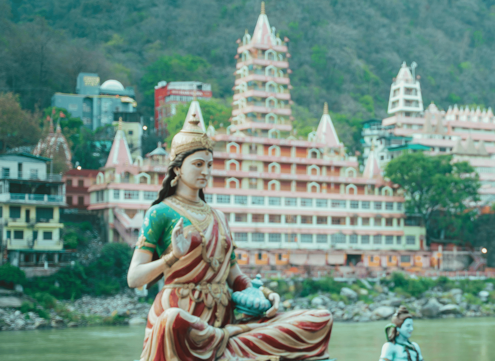 A woman practices yoga beside a river in Rishikesh, with a grand, multi-tiered ashram visible across the water.