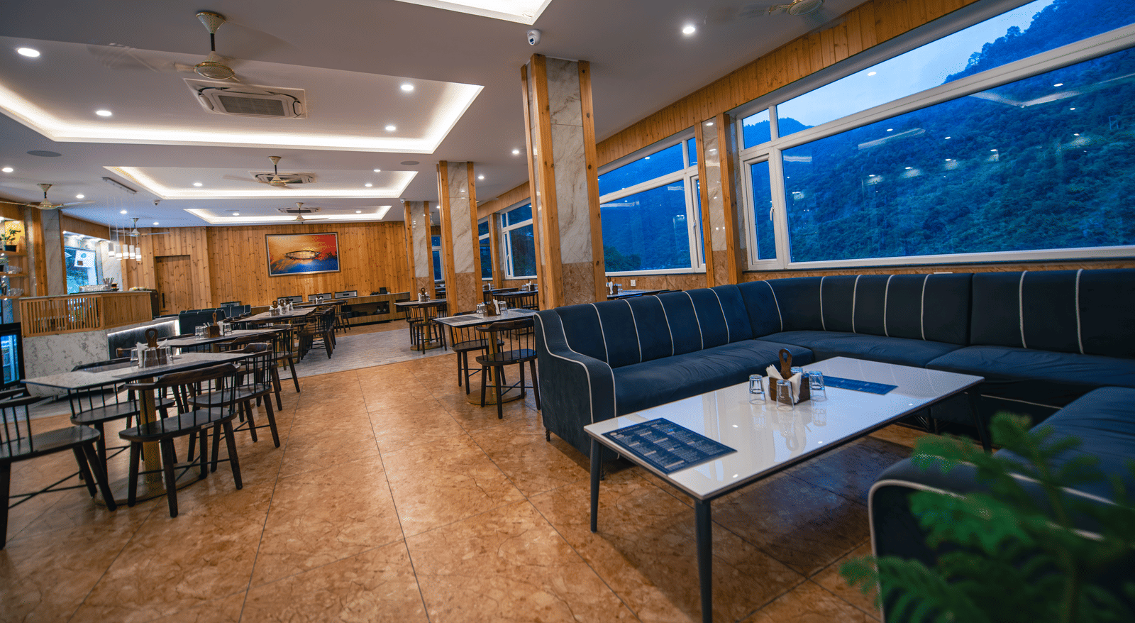 A restaurant at The Tattva Devaprayag with tables and blue chairs and a view of the mountains.