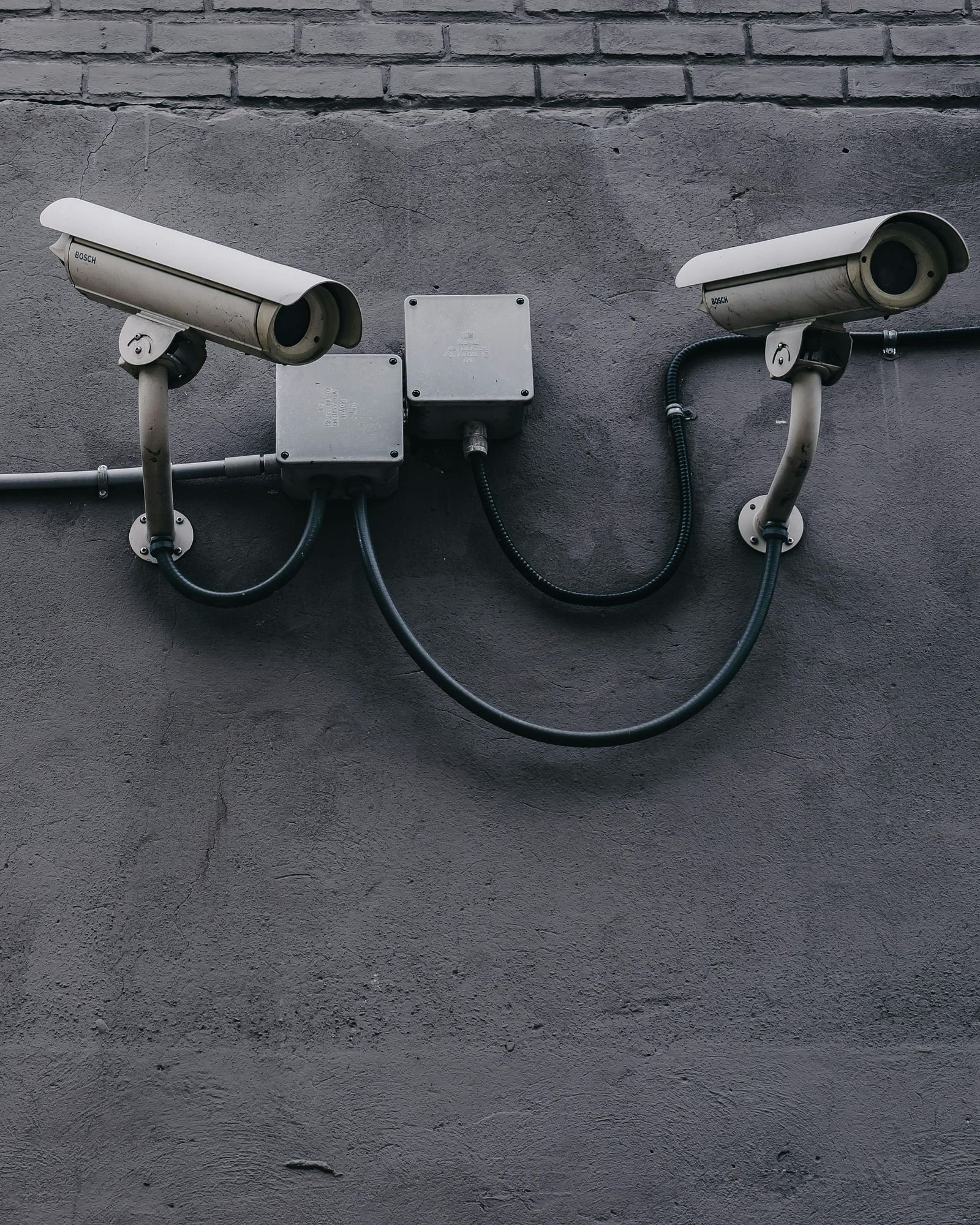 Two security cameras are mounted on a grey wall, connected by a black cable.