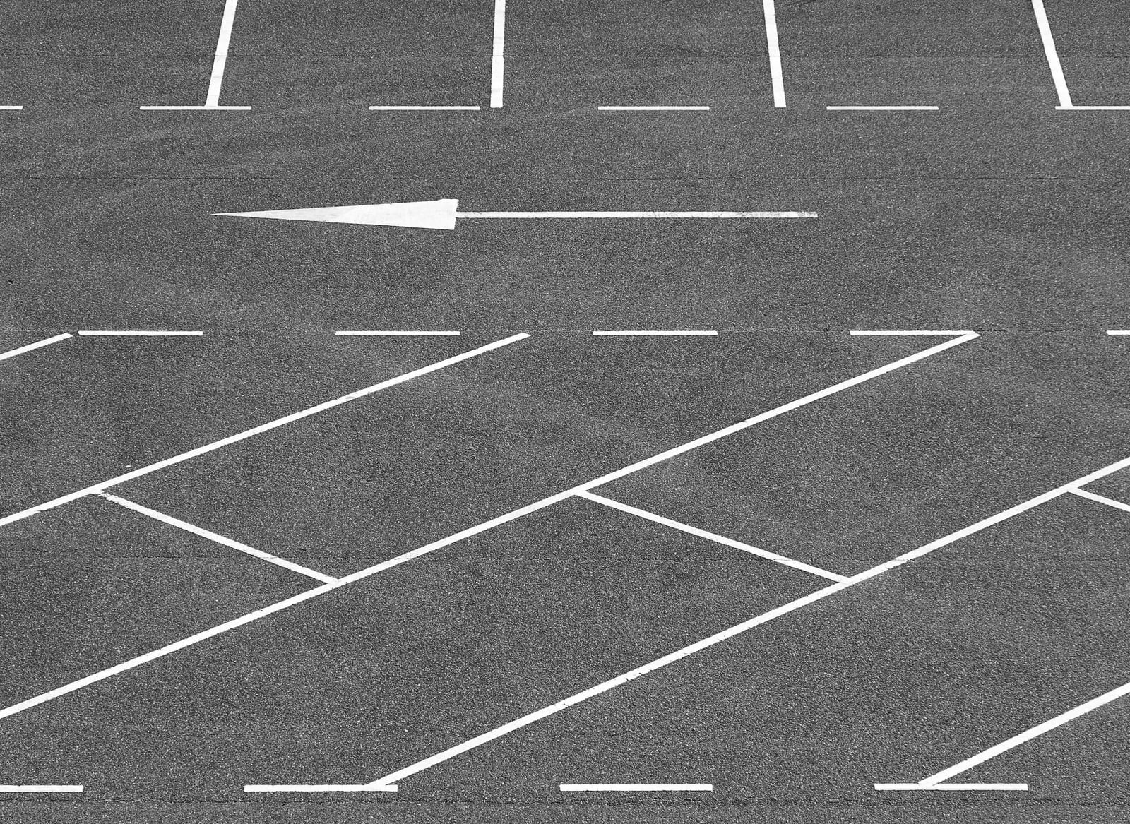 An empty asphalt parking lot is marked with white lines indicating parking spaces and a large arrow directing traffic flow.