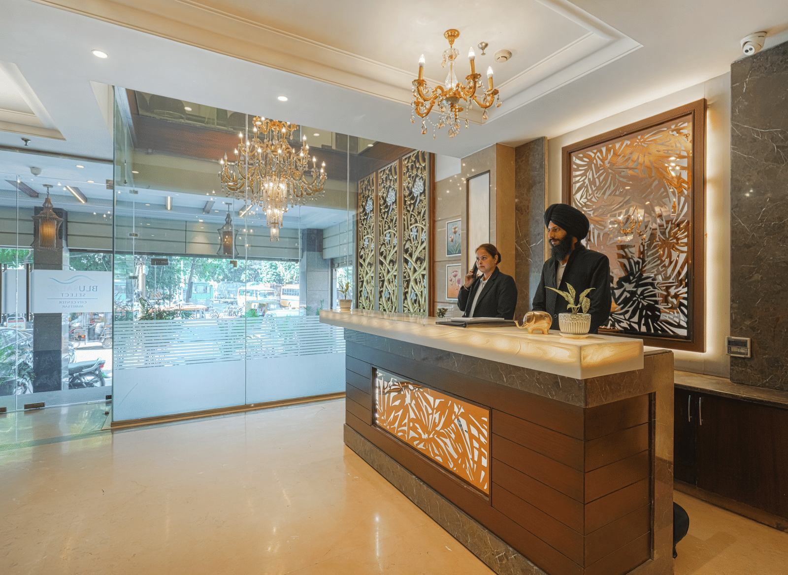 The hotel reception desk, staffed by a receptionist, viewed from the entrance through a glass partition at BluSalzz Select City Centre, Amritsar.