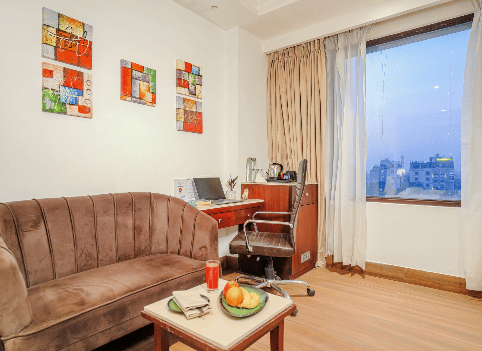 A small sitting and working area in a hotel room with a brown velvet sofa, a laptop on a desk, and colourful wall art at BluSalzz Select City Centre, Amritsar.