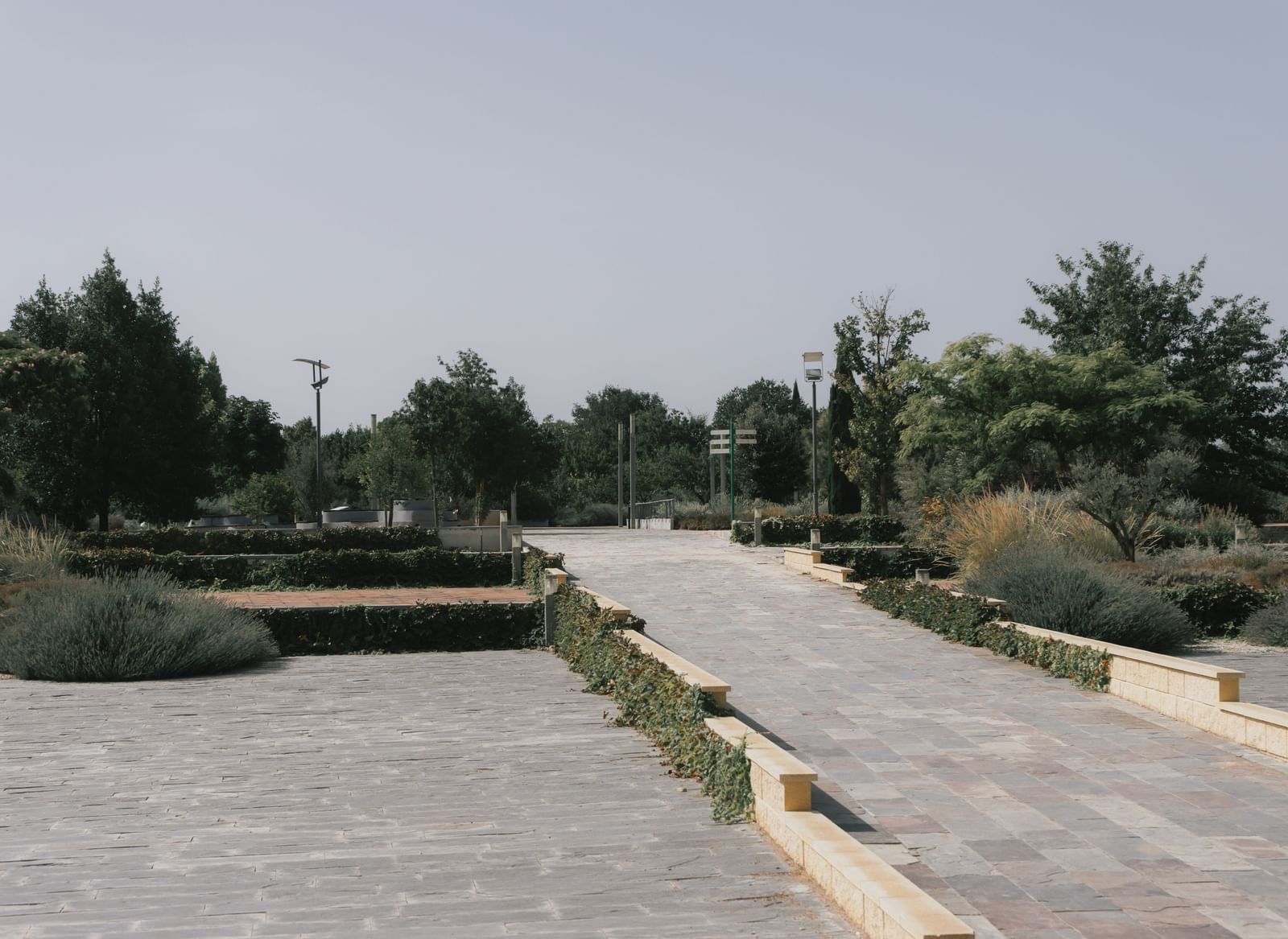 Wide, paved pathway through a formal garden or park under a bright, hazy sky.