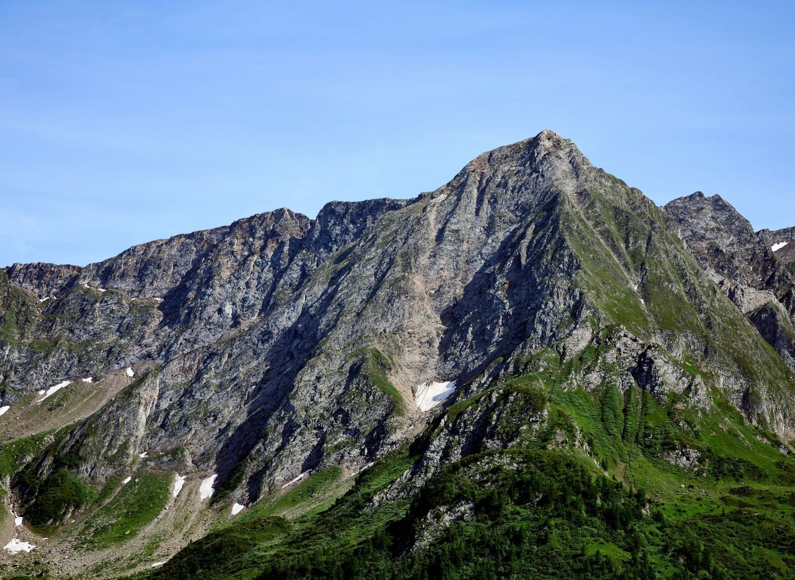A close-up view of a rugged mountain peak with patches of snow and green foliage under a clear blue sky.