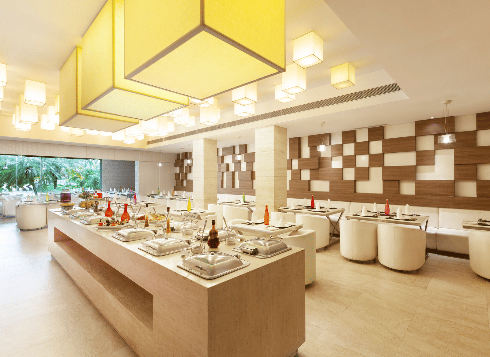 A diagonal view of a restaurant with an island in the middle featuring buffet items, surrounded by tables and chairs on the sides | Cenneys Gateway