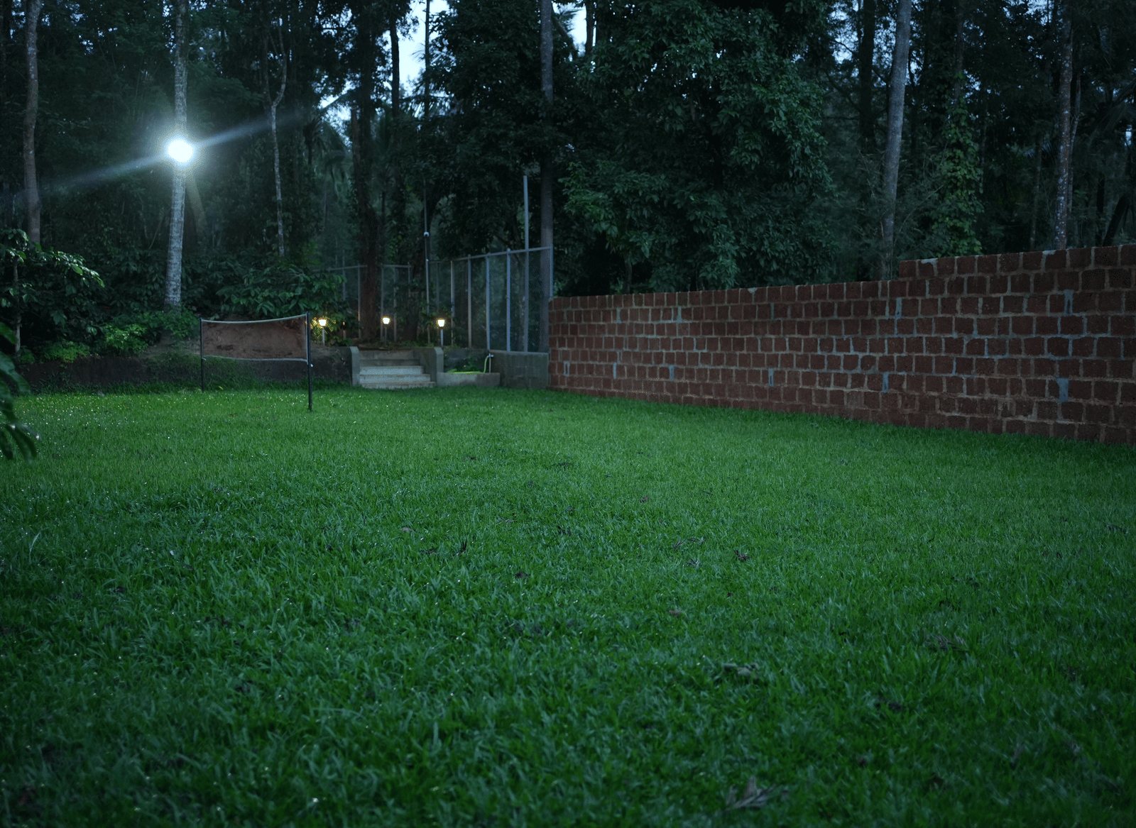 A well-kept grassy lawn bordered by a low brick wall and dark trees, lit by a single lamp at night at Coorg Orange Blossom Resort and Spa.