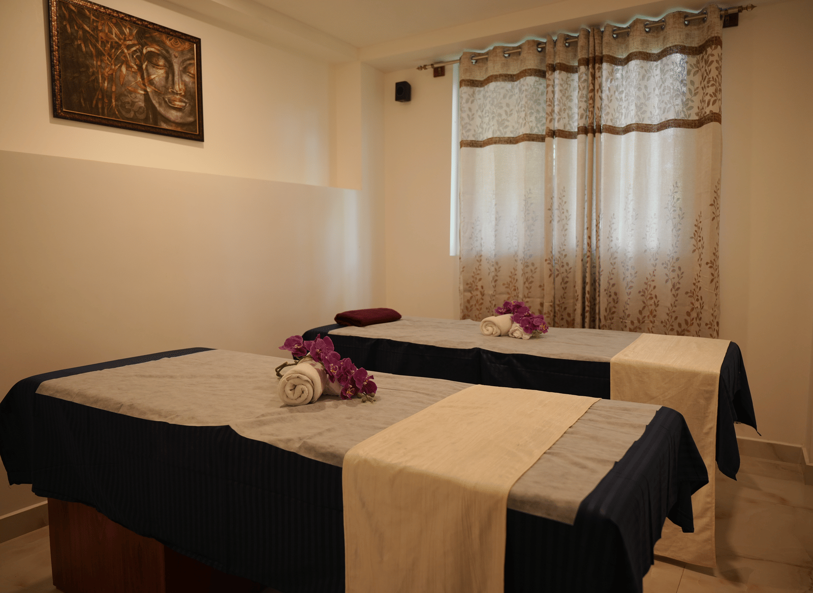 A spa treatment room containing 2 massage tables, sheer curtains, and simple wall decor at Coorg Orange Blossom Resort and Spa.