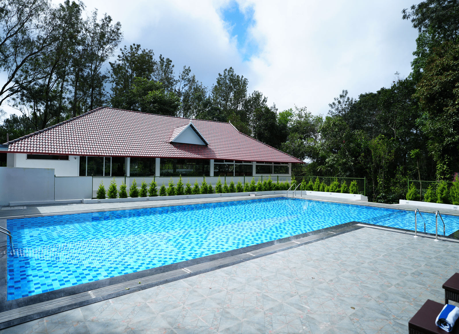 An outdoor swimming pool next to a white and red-roofed building, surrounded by paved decking and greenery at Coorg Orange Blossom Resort and Spa.