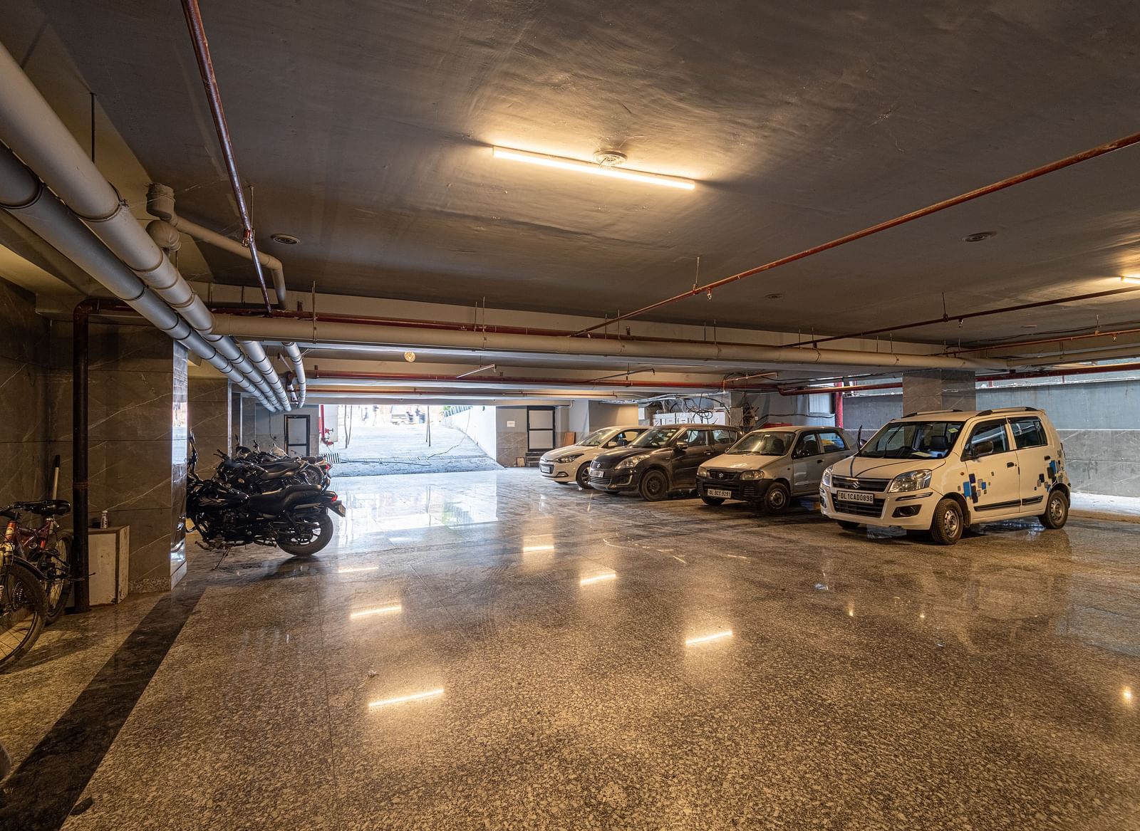 Image of a large parking space at DLS Grand Luxotica, Dehradun featuring cars and bikes parked in warm lighting