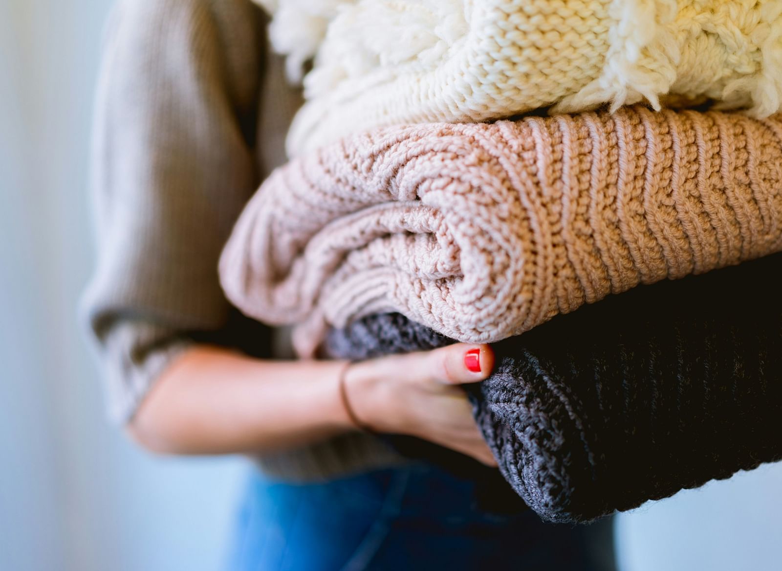 A person holding a stack of soft, folded sweaters in various neutral colours.