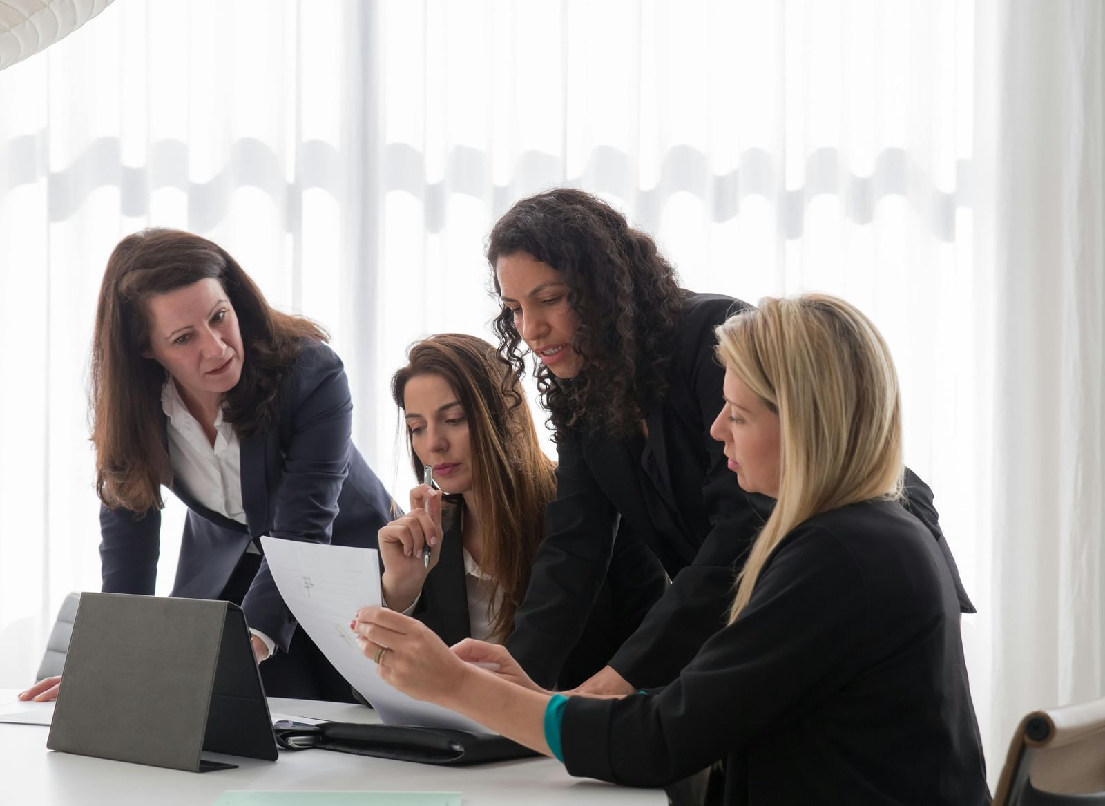 Four businesswomen in blazers are gathered around a table in an office setting, looking at document and engaged in discussion. 