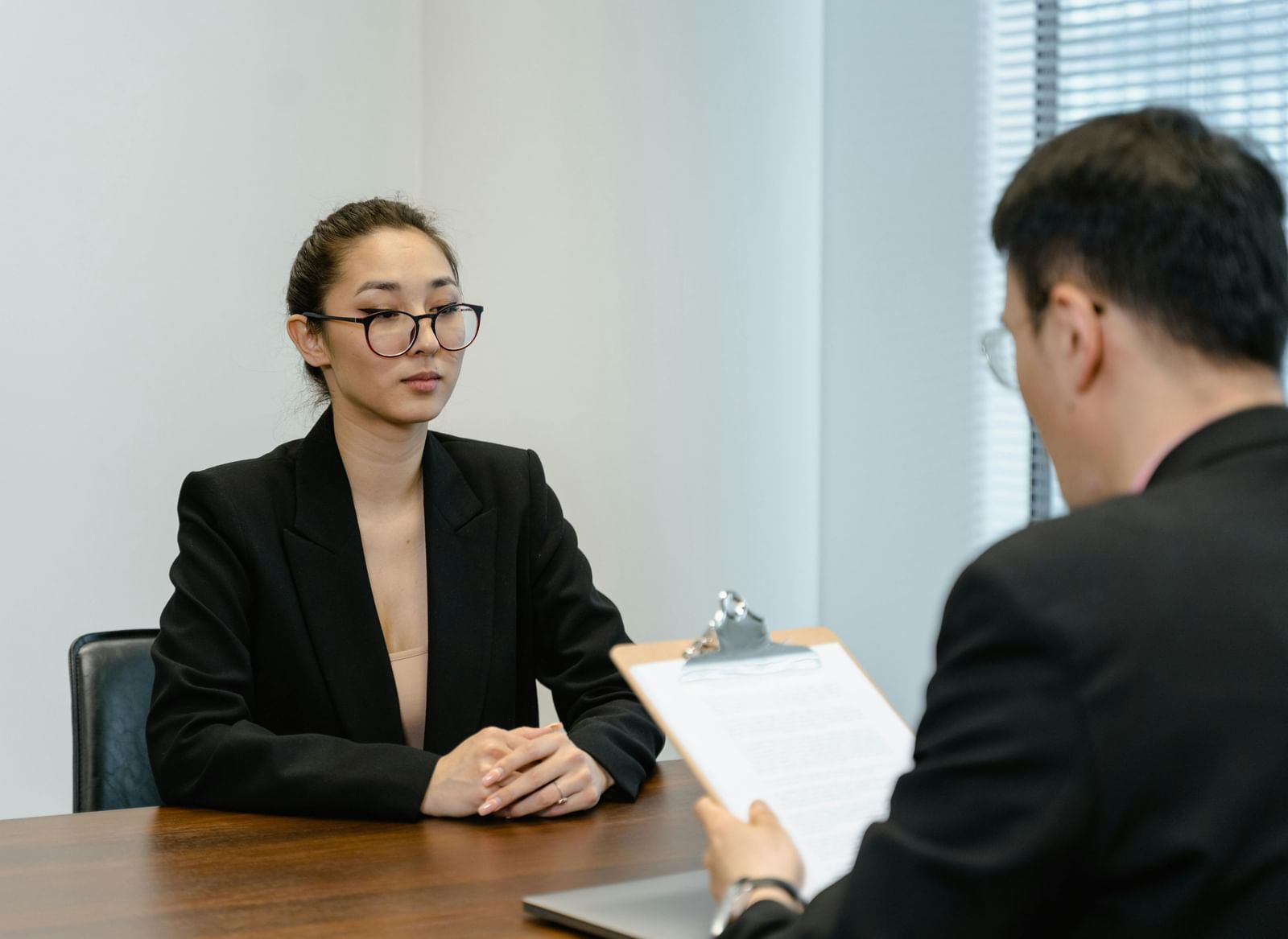 Two professionals with black suits are engaged in a  interview at an office desk with a clipboard. 