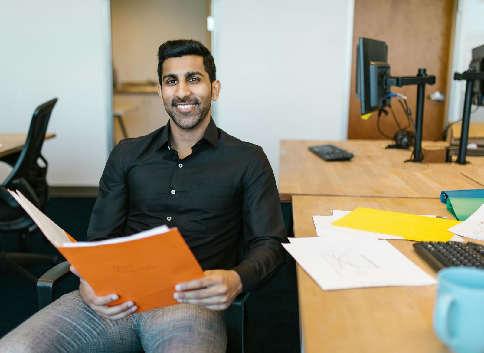 A smiling man in a black shirt holds an orange folder in an office setting. 