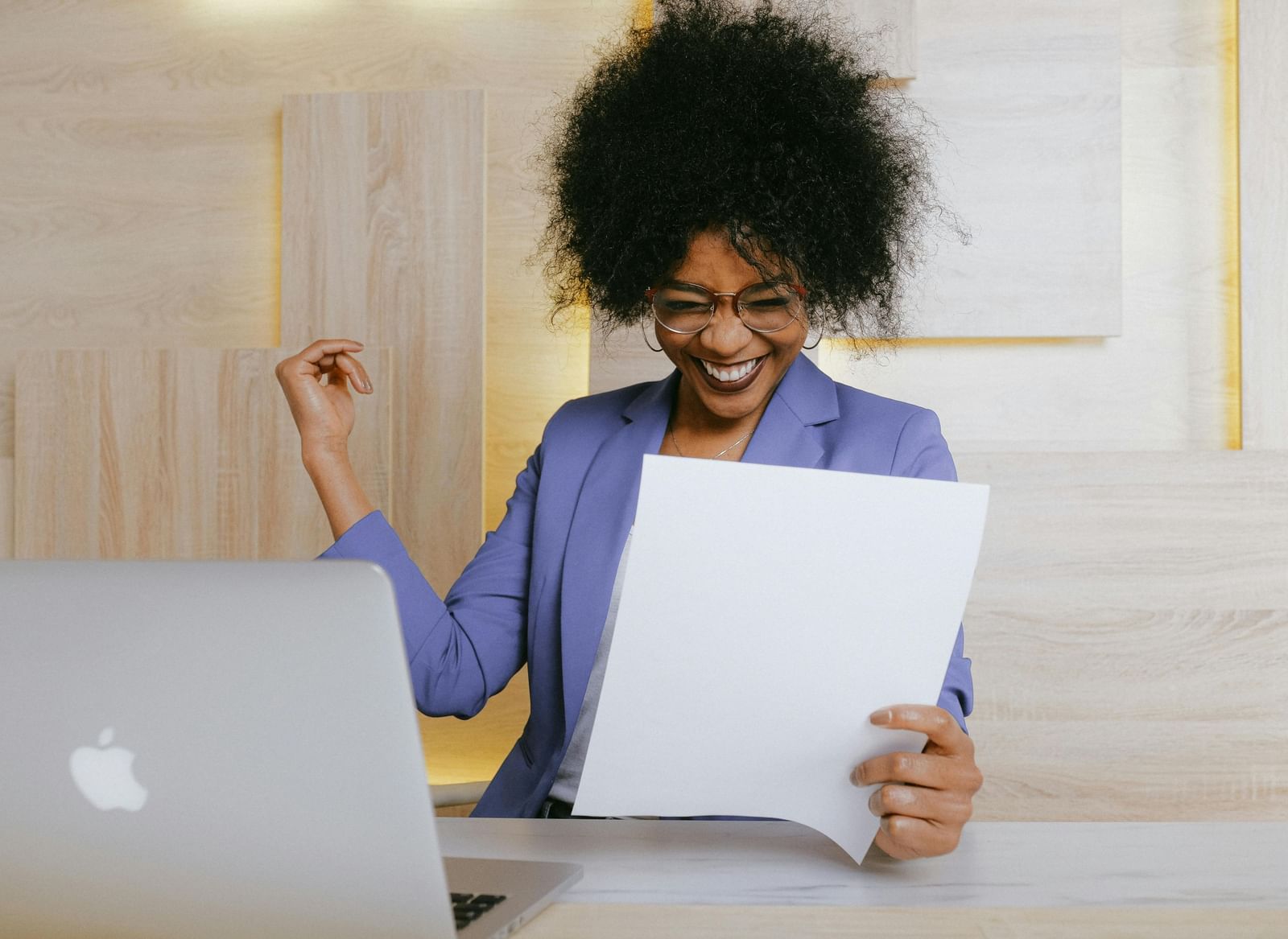 A woman with dark, voluminous hair, wearing a purple blazer, smiles broadly while holding a sheet of paper, with a laptop visible on a desk in front of her.