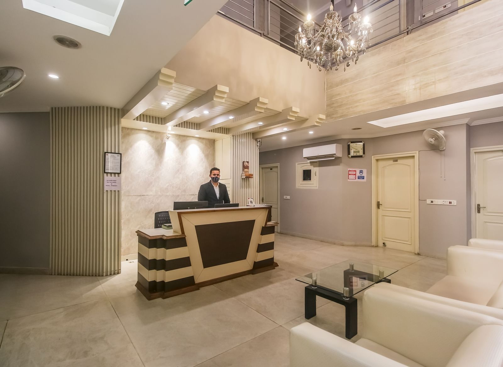 A hotel lobby with a reception desk, a chandelier, a seating area with white sofas, and a staff member behind the counter, lit by soft lighting. - Hotel Diamond Plaza, Chandigarh.