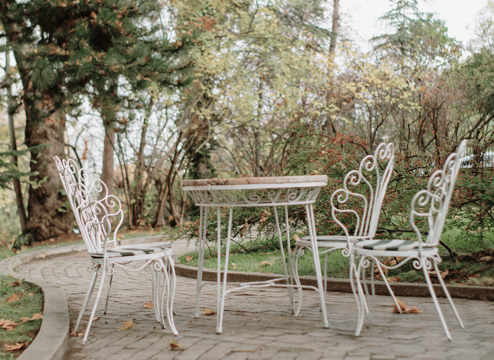 An outdoor patio setup featuring a round table and 2 matching ornate chairs arranged on a paved surface surrounded by trees and bushes.