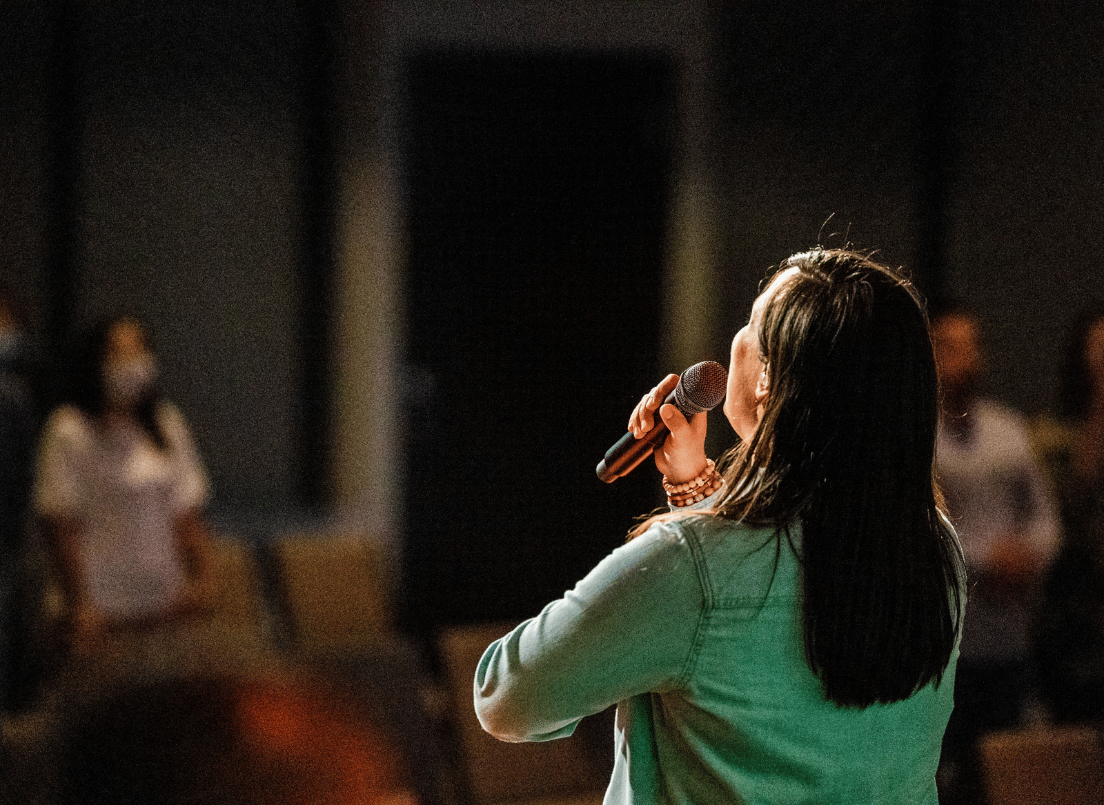 A person speaking or singing into a microphone in front of an audience in a dimly lit room.