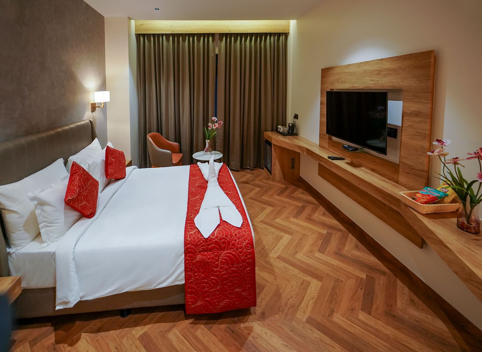 A hotel room features a bed with pillows and folded linens, curtains, a mounted television, and a long desk area with flowers - Hotel Park Inn, Nellore.