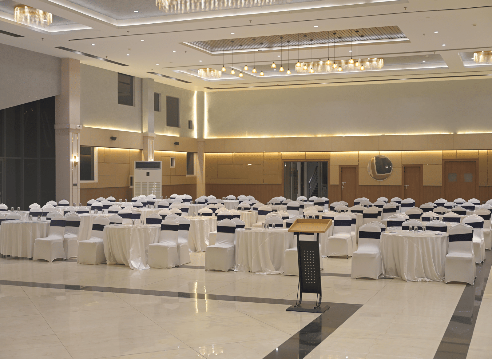 A front-angled view of the banquet hall set with round tables and white seating under warm ceiling lights at Hotel Sonar Bangla Mayapur.