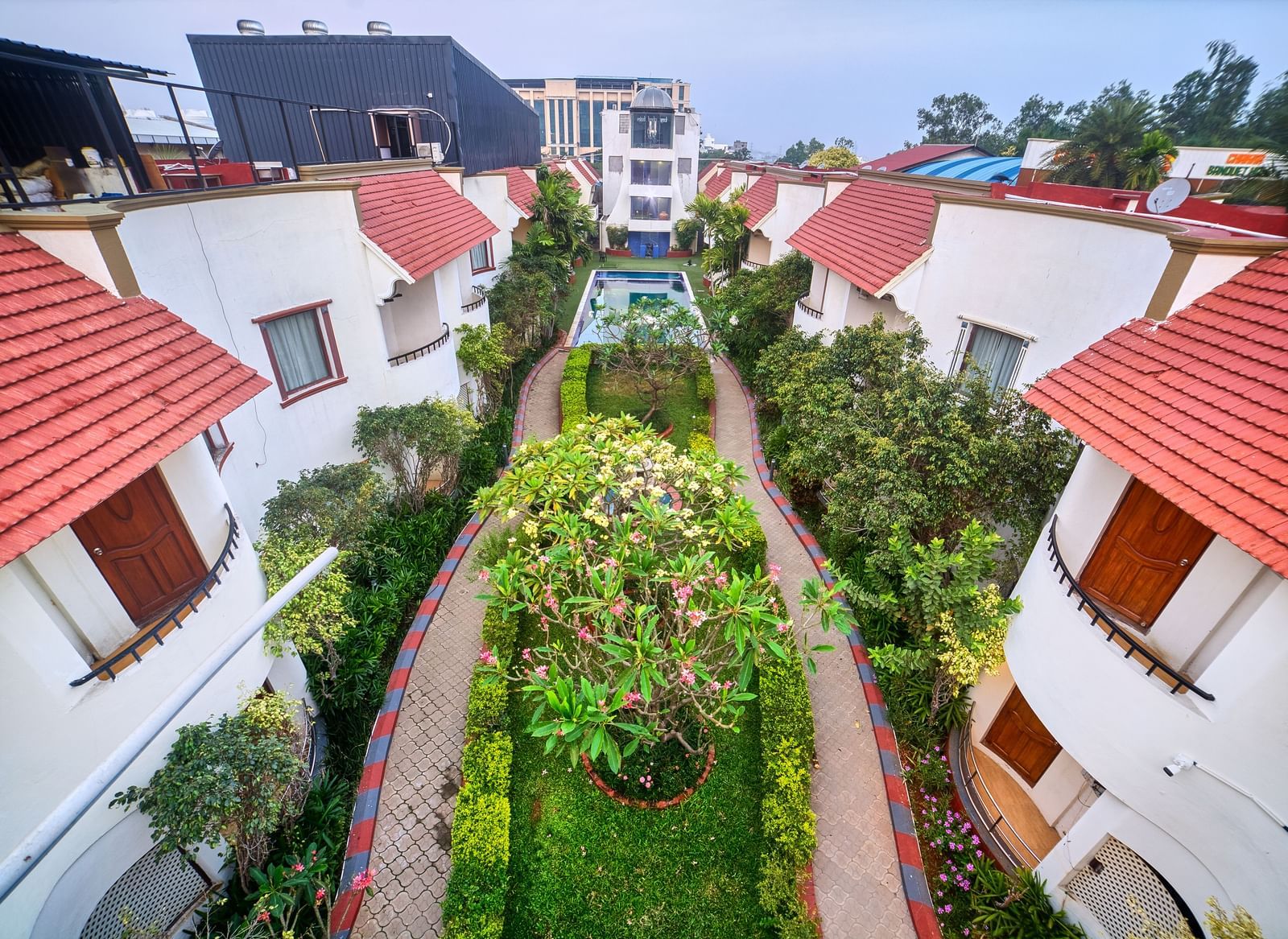 Elevated view showcasing a row of villa-style accommodations with striking red tiled roofs and lush green pathways between them at Ilara Hotels & Spa, OMR, Chennai.