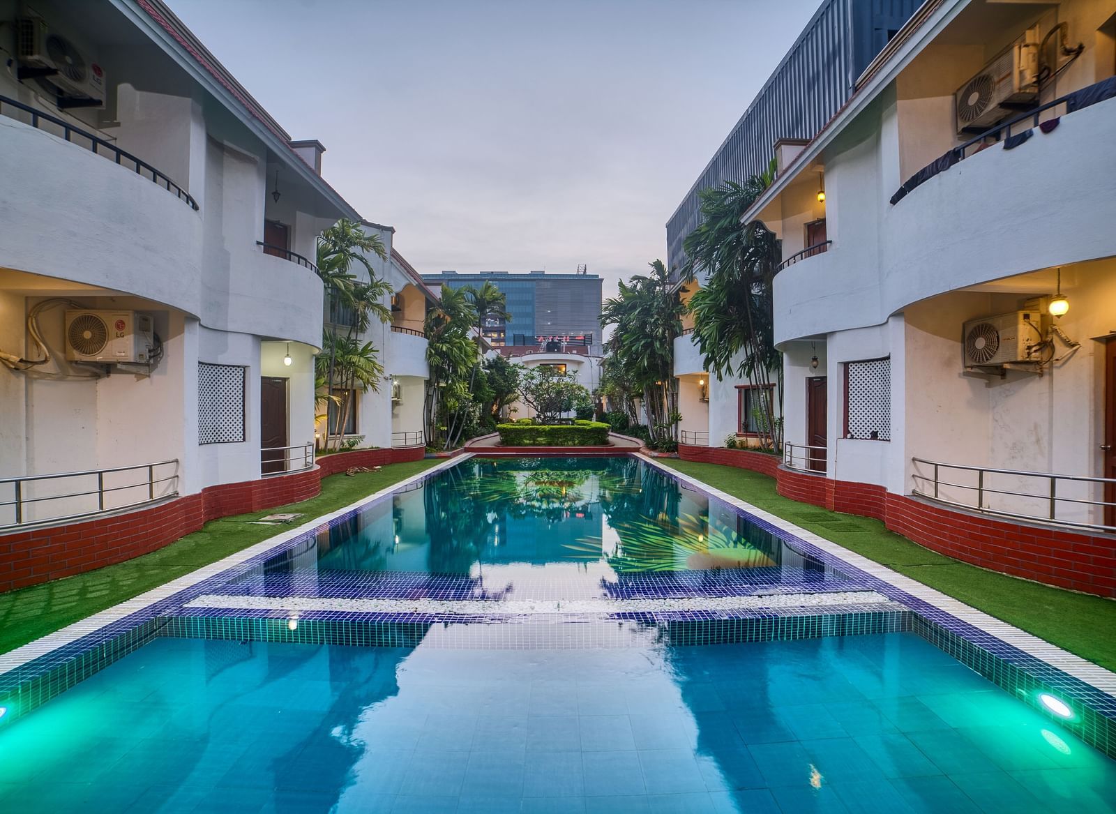 A symmetrical view of Ilara Hotels & Spa, OMR, Chennai illuminated at dusk, lining a long, reflective swimming pool or water feature.