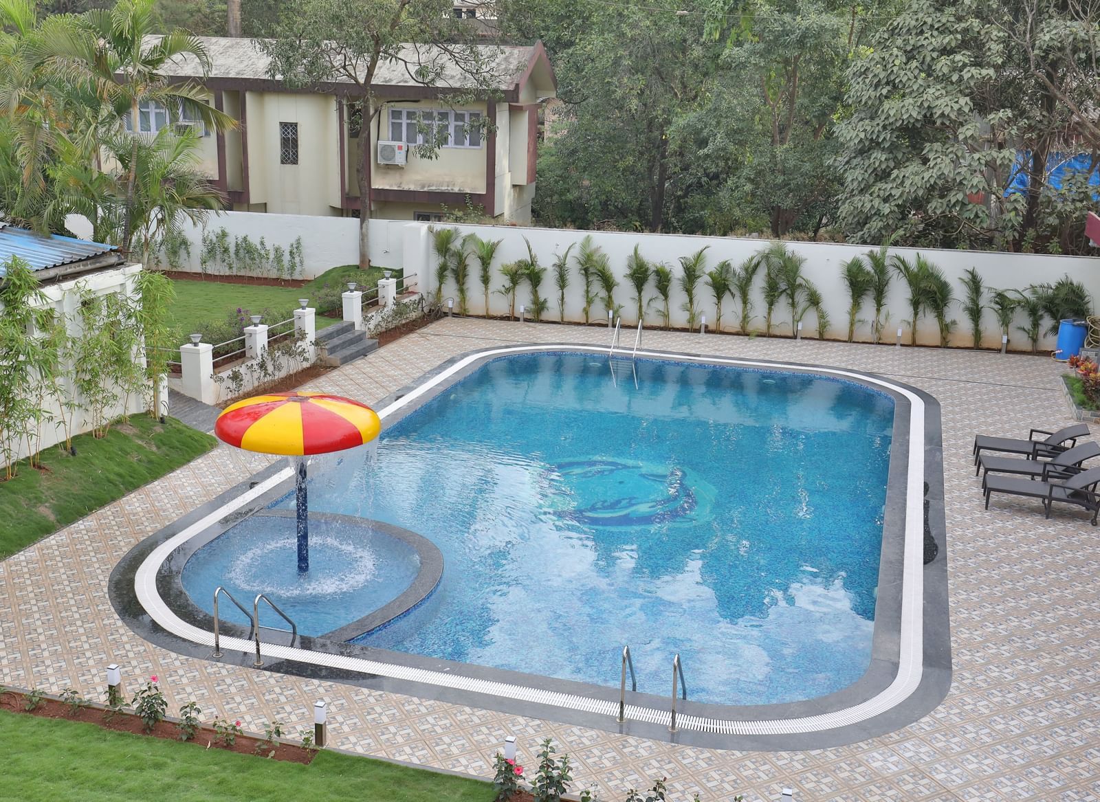 An outdoor swimming pool at Imperial Grande during the day, with an umbrella-shaped water feature, surrounded by paved areas and some greenery.