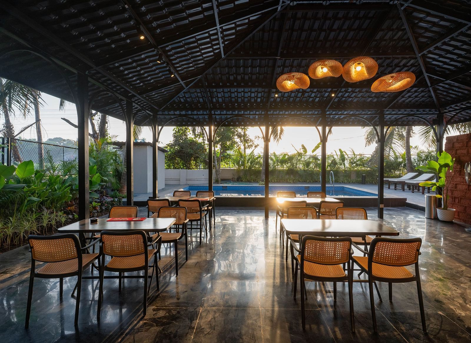 An outdoor dining area under a dark, peaked roof, open to the surrounding scenery. Several wooden tables and chairs are neatly arranged, with a glimpse of a swimming pool visible in the background under the bright sky at Kssemya