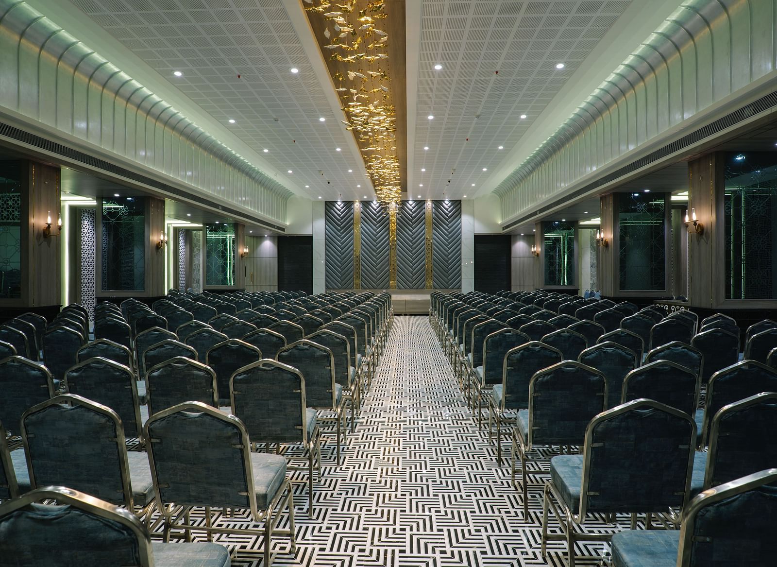 A large room is set up with rows of dark chairs facing a stage or presentation area - Melior Inn