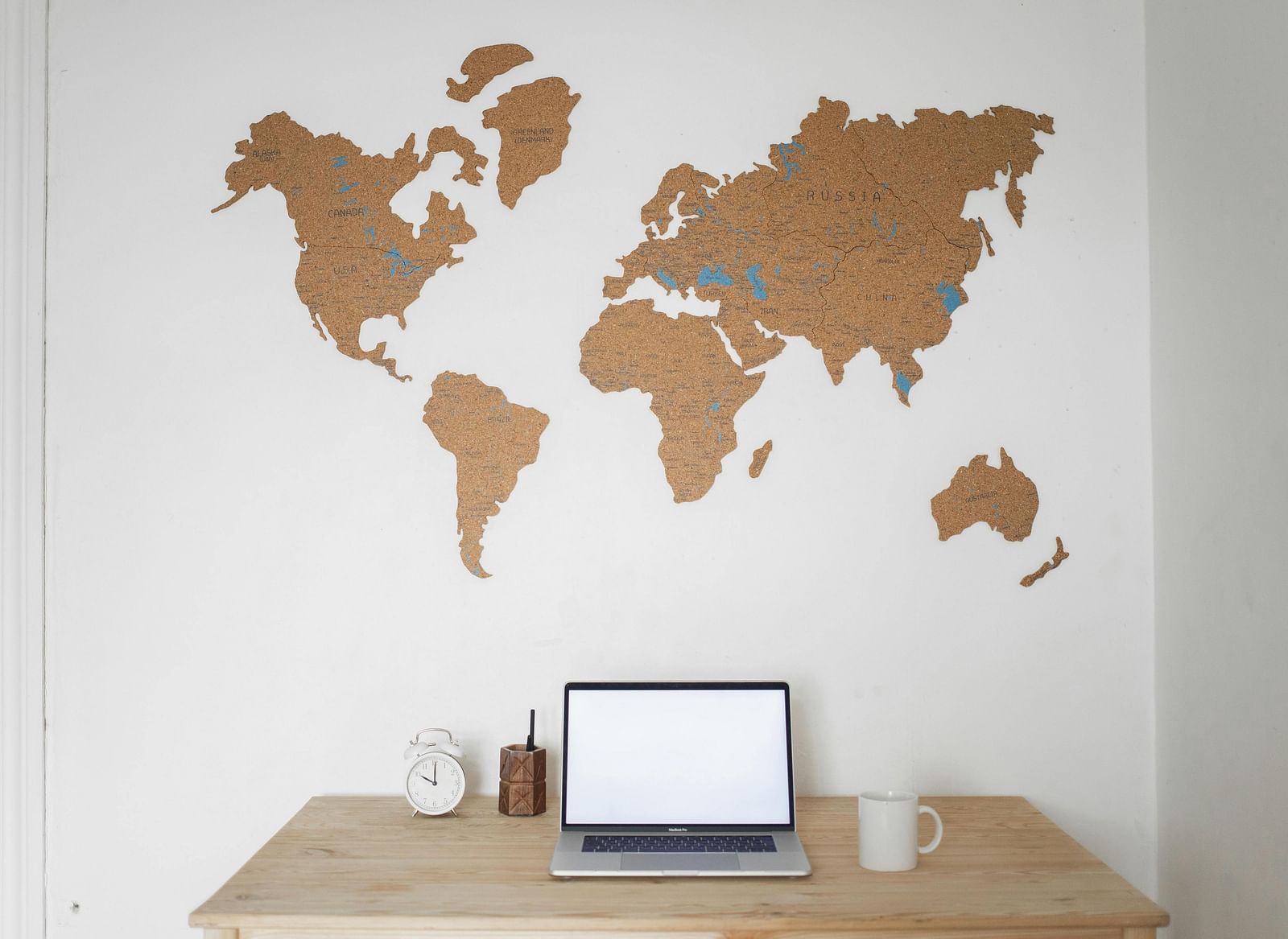 A laptop with a blank white screen sits on a desk in front of a wooden world map on a white wall.