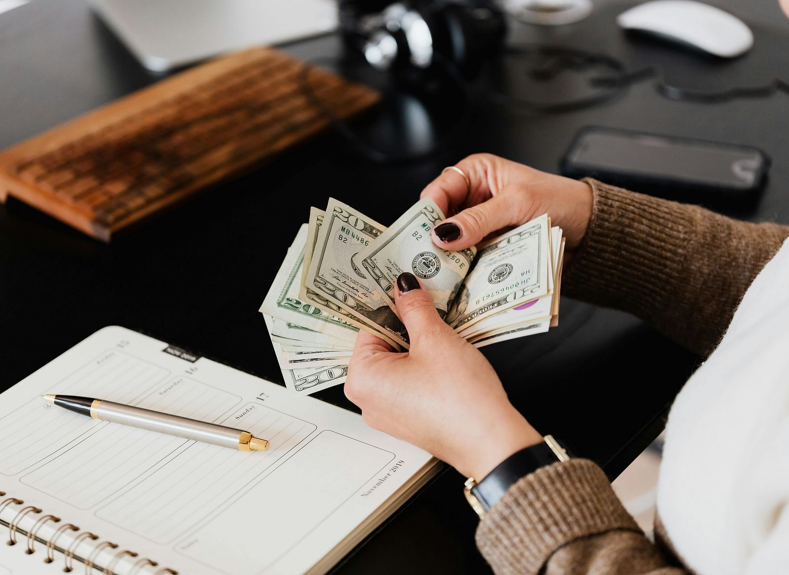 A person holding a stack of currency notes over an open planner on a black desk.