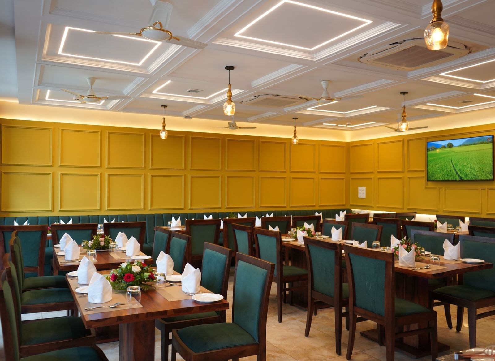 A wide shot of a dining hall with multiple tables set for a meal. The chairs are dark green, and the walls are yellow. A TV is visible on the far wall.