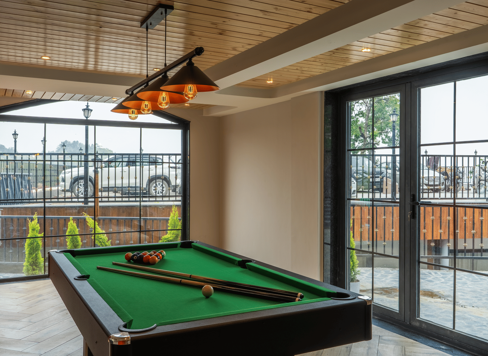 A green billiard table with cues, inside a room with large glass windows and hanging lights at Perfectstayz Luxe Mall Road.