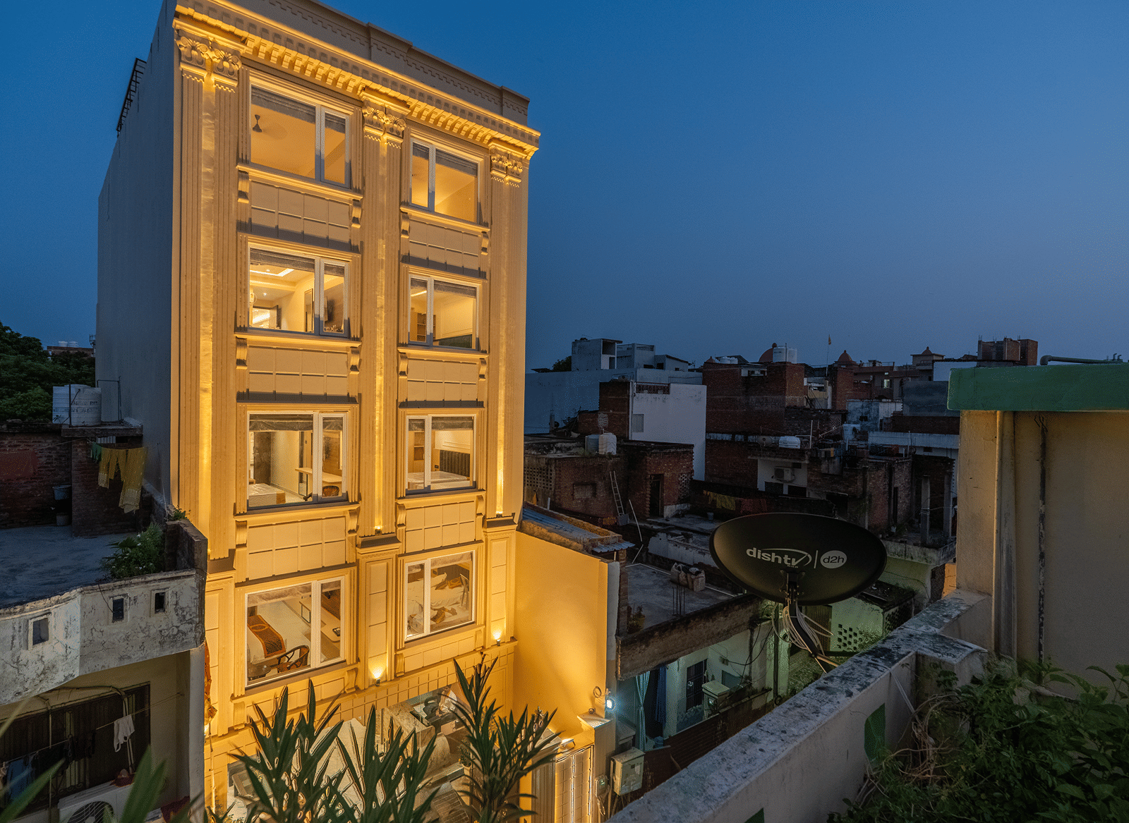 Angled evening view of the lit cream facade of Perfectstayz Premium Lavender, Varanasi, seen from a neighbouring rooftop with a satellite dish and city skyline.
