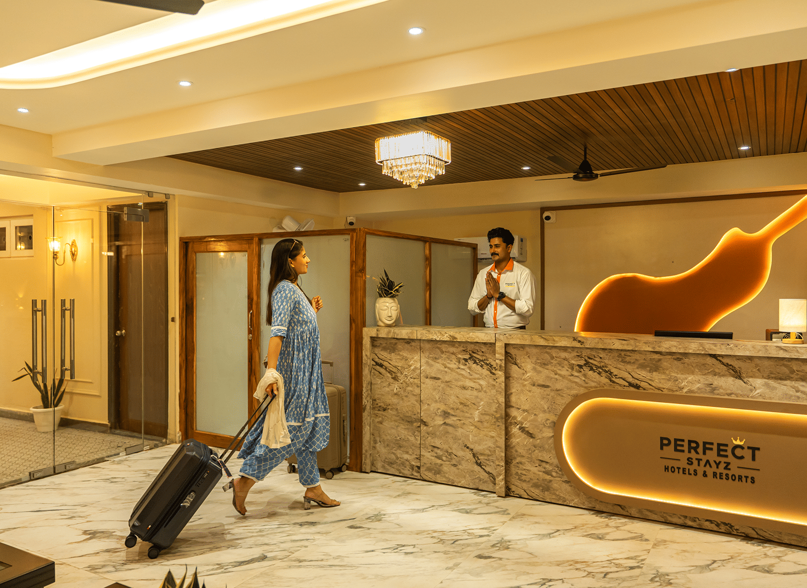 Guest with a suitcase arriving at the reception of Perfectstayz Premium Lavender, Varanasi, greeted by a staff member behind a marble desk with a backlit logo.
