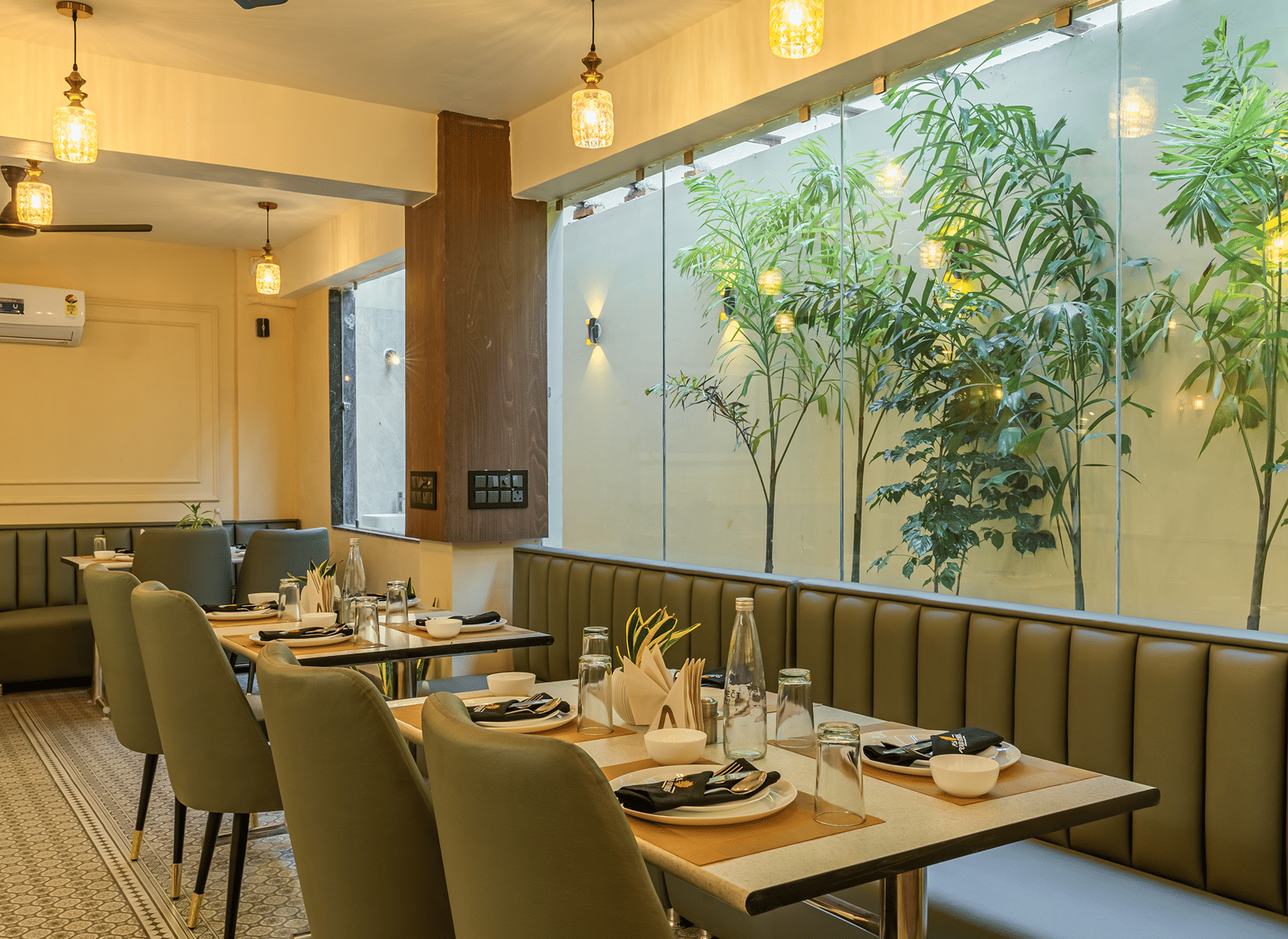 Set dining table at the restaurant of Perfectstayz Premium Lavender, Varanasi with plates, glasses, and a water bottle beside green banquette seating and plants.