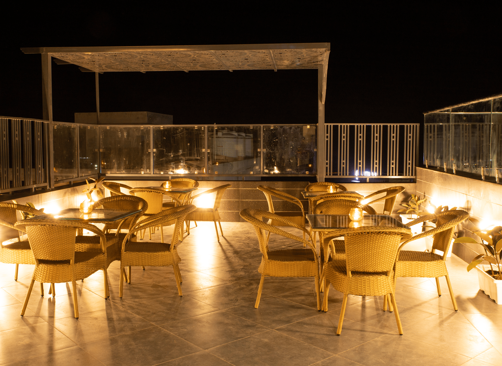 An outdoor rooftop cafe at Perfectstayz Premium Laxmi Heritage with woven chairs and tables on a patio, warmly lit under a dark sky, enclosed by a glass railing.