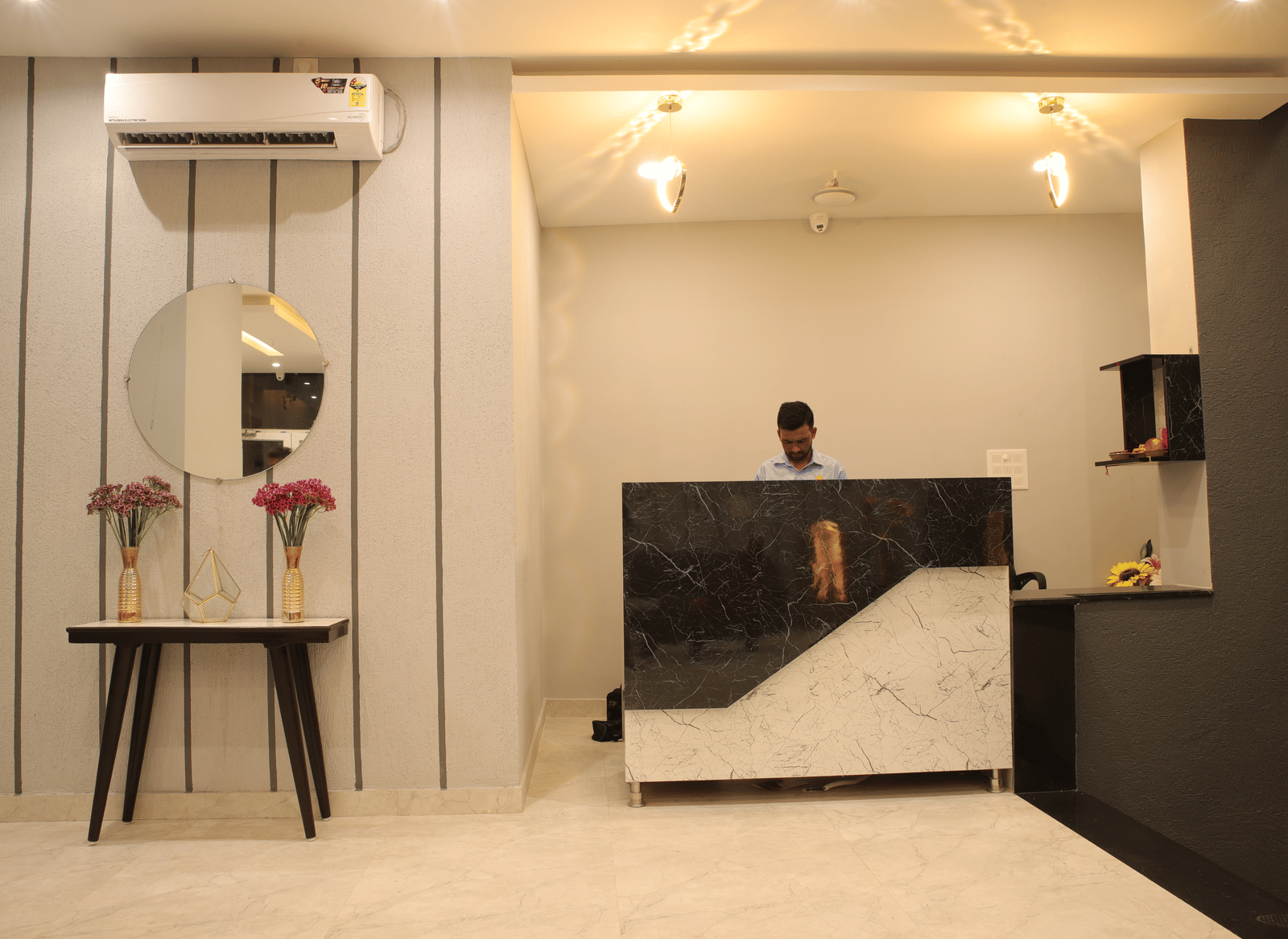 A man standing behind a reception desk at Perfectstayz Premium Laxmi Heritage, with light walls, ceiling lights, a mirror, flower vases, and an air conditioner above.